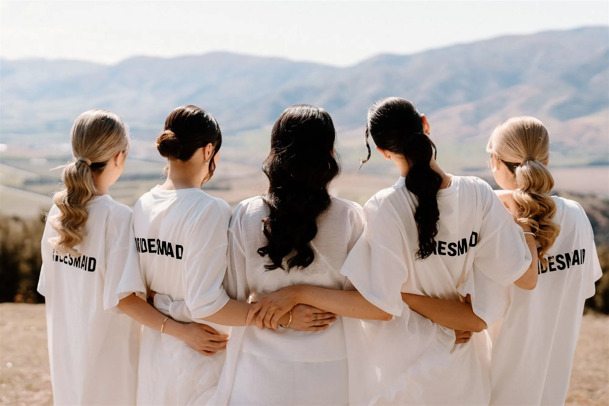 Five bridesmaids from behind, arms linked, with curled ponytails and low buns